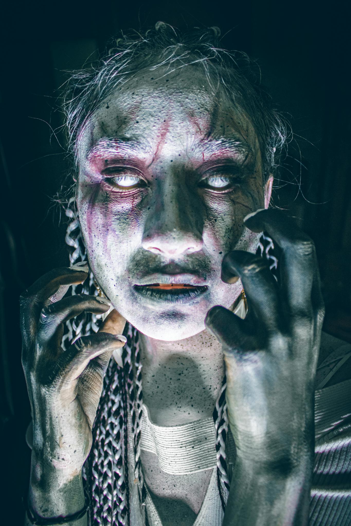 Eerie figure with ghostly makeup and intense expression in dark ambience.